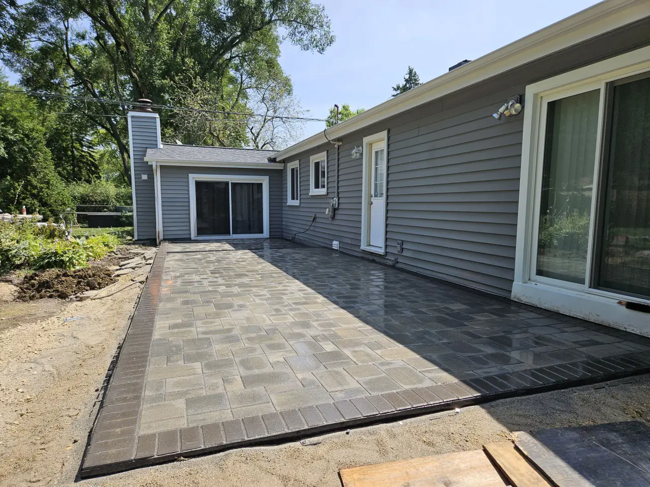 Backyard paver patio installation with gray concrete pavers and dark border edging along residential home exterior